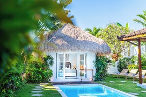 Un bungalow au toit de chaume, doté d’une piscine, entouré d’une végétation luxuriante et surplombant un ciel dégagé.