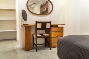 Une pièce avec un bureau en bois, une chaise dont le dossier est en osier tressé, un miroir au mur et un lit recouvert d’une couverture grise.
