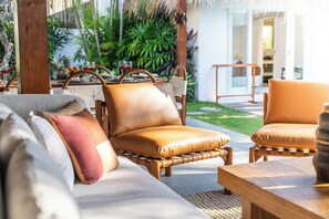 A cozy outdoor seating area with wicker chairs and a wooden table, surrounded by lush greenery and a modern building in the background. - Villa Thiara Oberoi Seminyak 500m Kudeta beach (Seminyak)