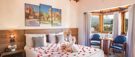 A hotel room with a large bed, a nightstand, a bedside table, a chair, a small table, and a window with curtains.