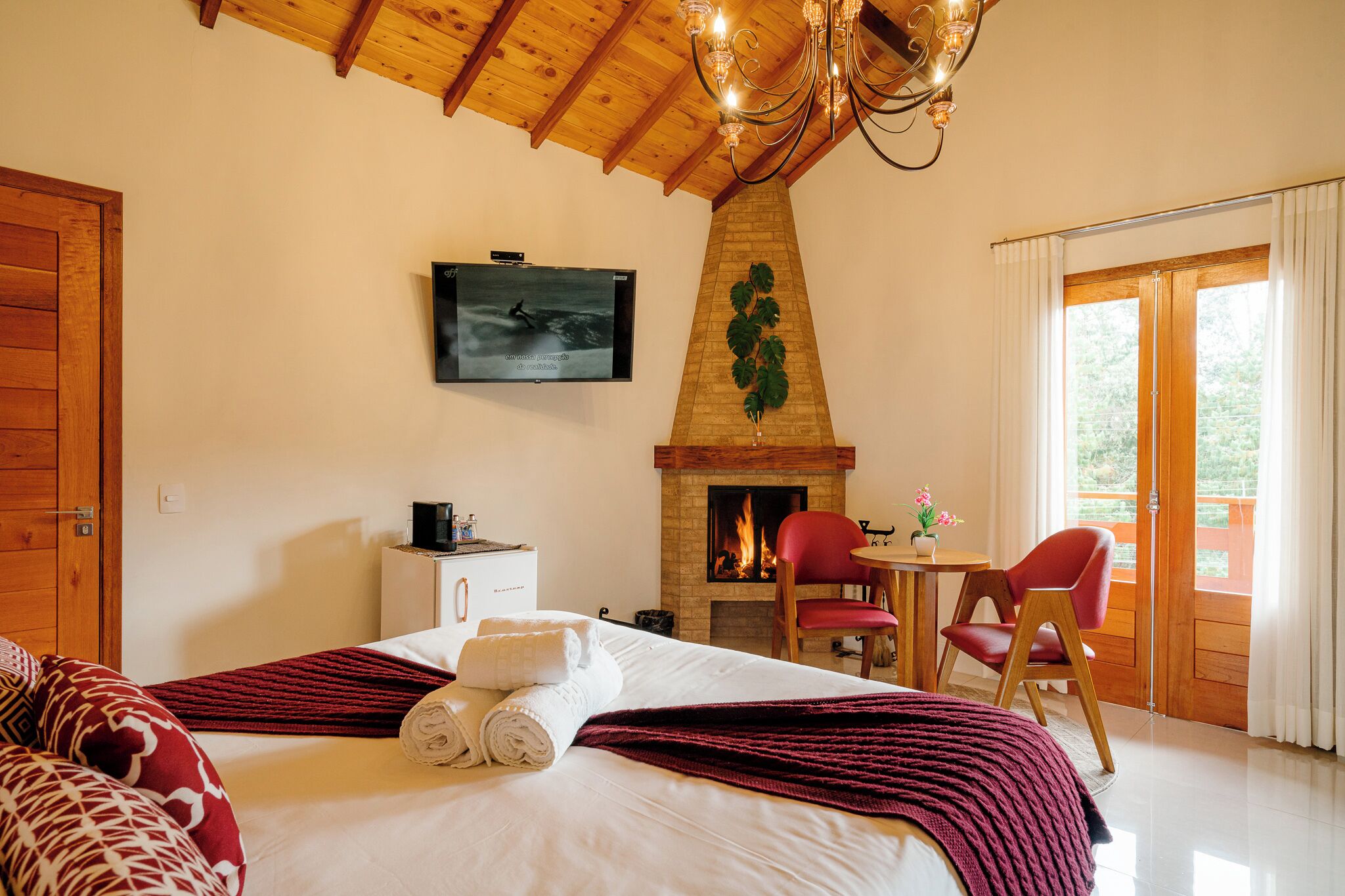 A hotel room with a bed, a television, a fireplace, a dining area, and a view through a glass door.