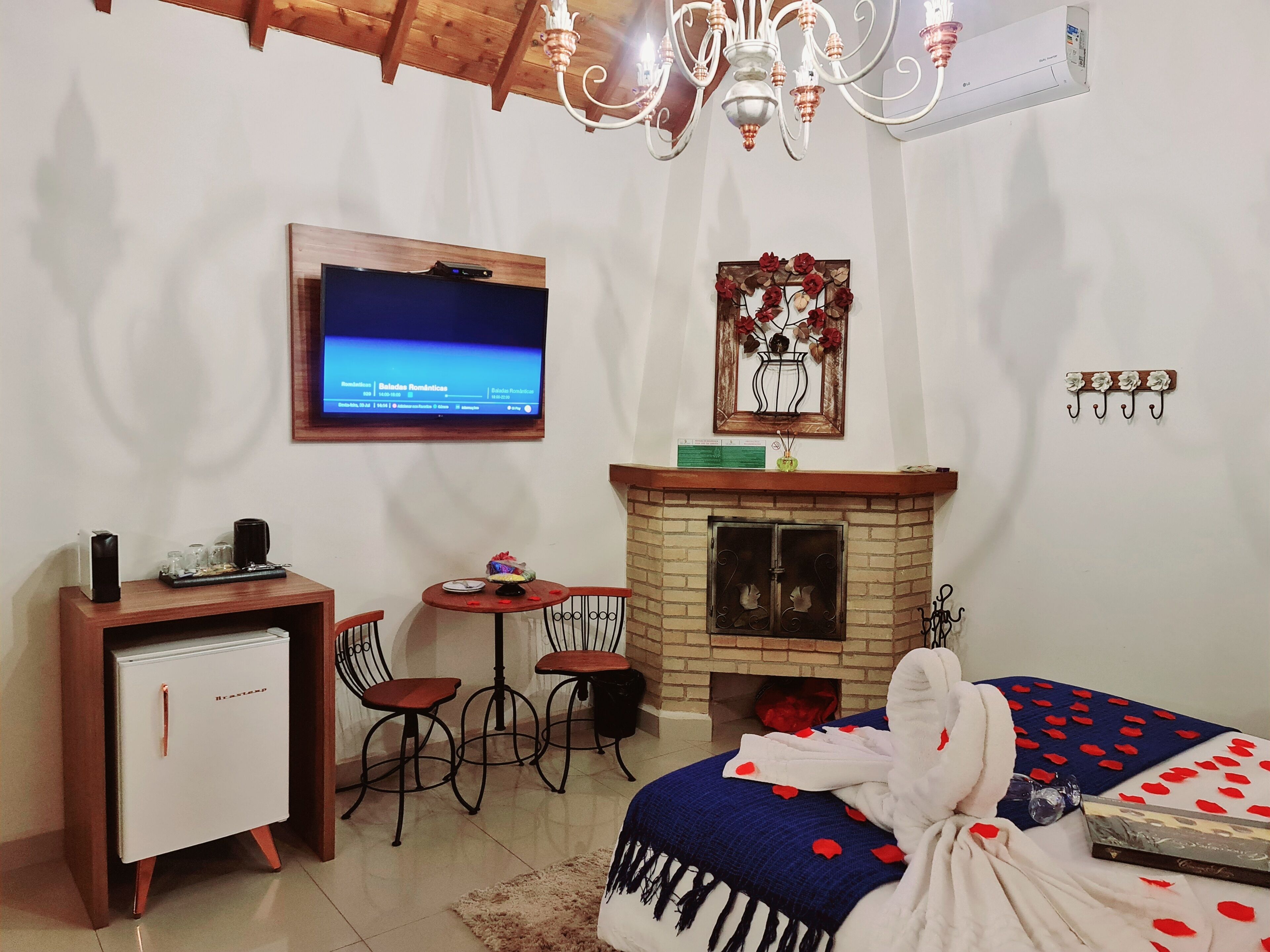 A cozy bedroom with a fireplace, a TV mounted on the wall, a desk with a computer, and a bed with a rose-patterned blanket.