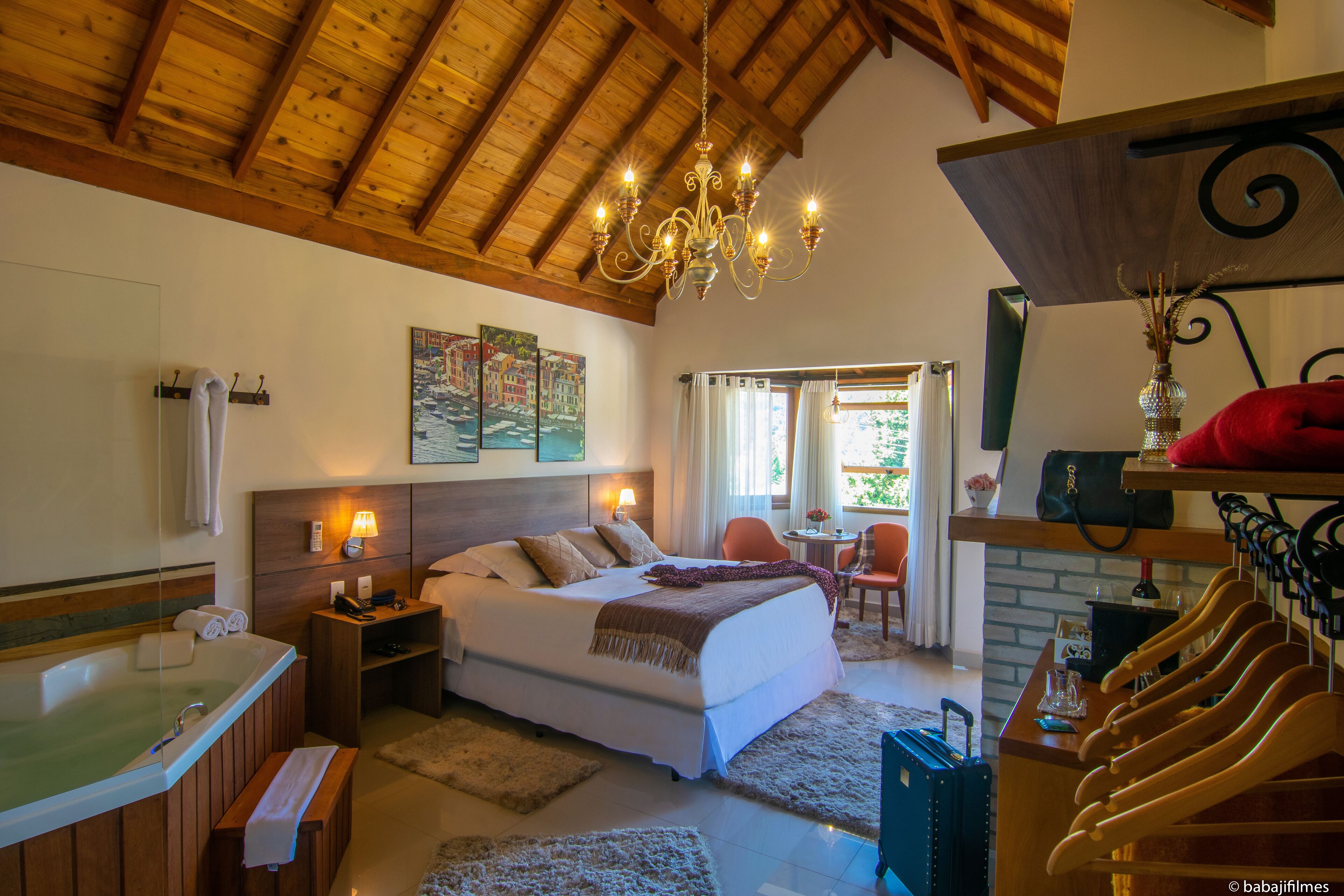 A bedroom with a large bed, a wooden ceiling, a chandelier, a bathroom with a bathtub, and a seating area with a table and chairs.
