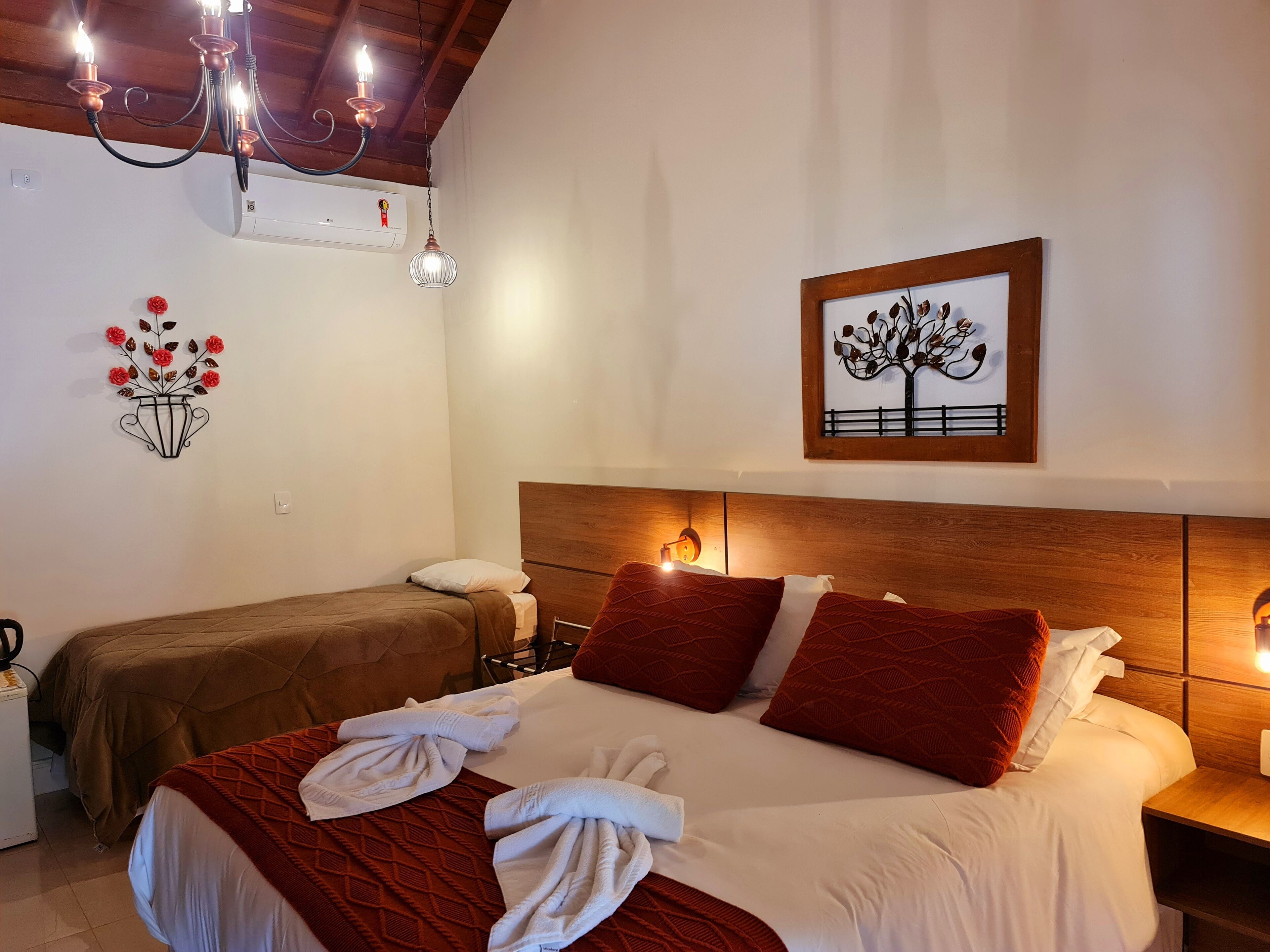 A hotel room with a wooden headboard, a bed with red and white bedding, a nightstand with a lamp, a wall-mounted air conditioner, and a decorative wall piece.