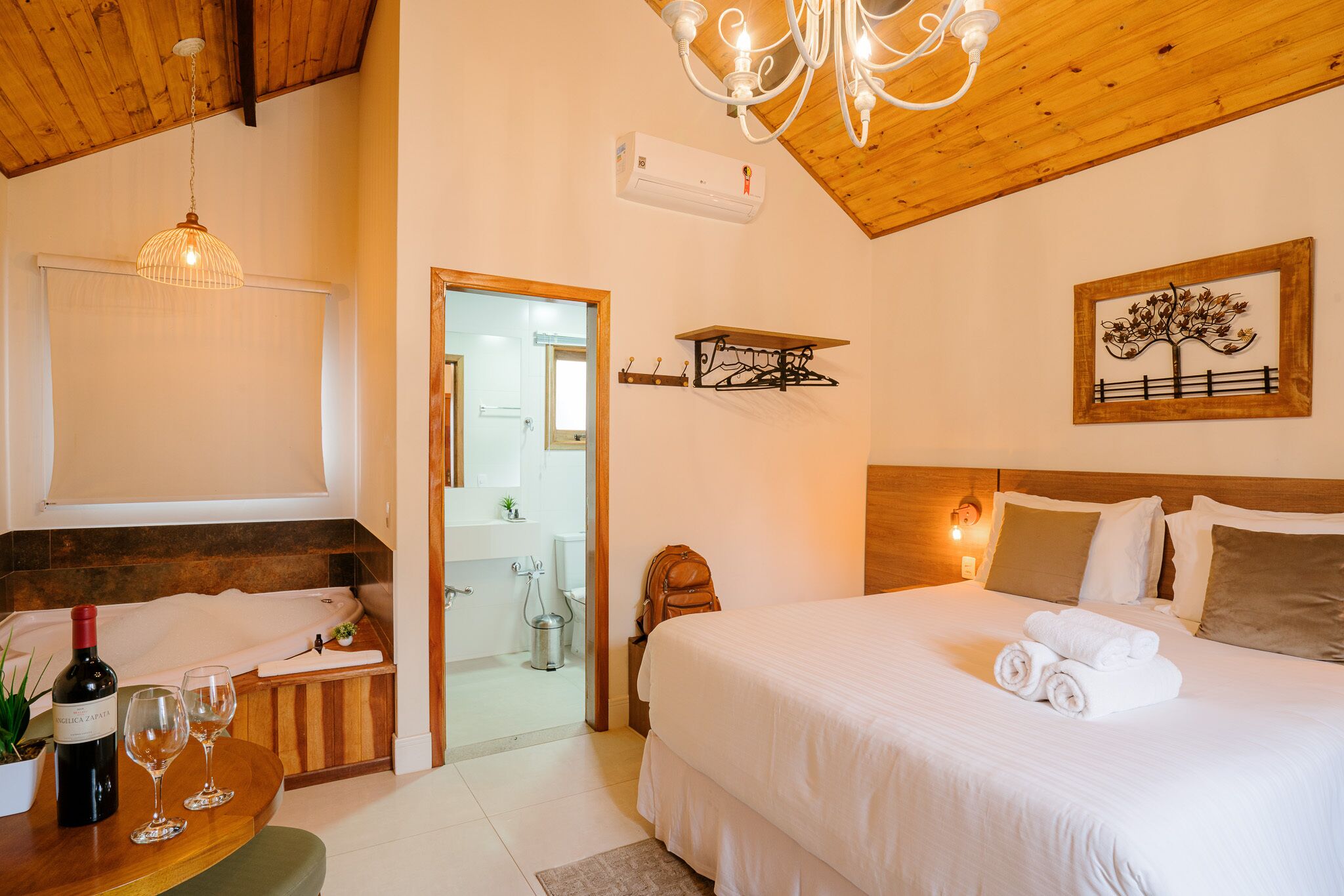 A hotel room with a wooden ceiling, a chandelier, a bed with white linens, a small table with a wine bottle and glasses, and a bathroom visible through an open door.