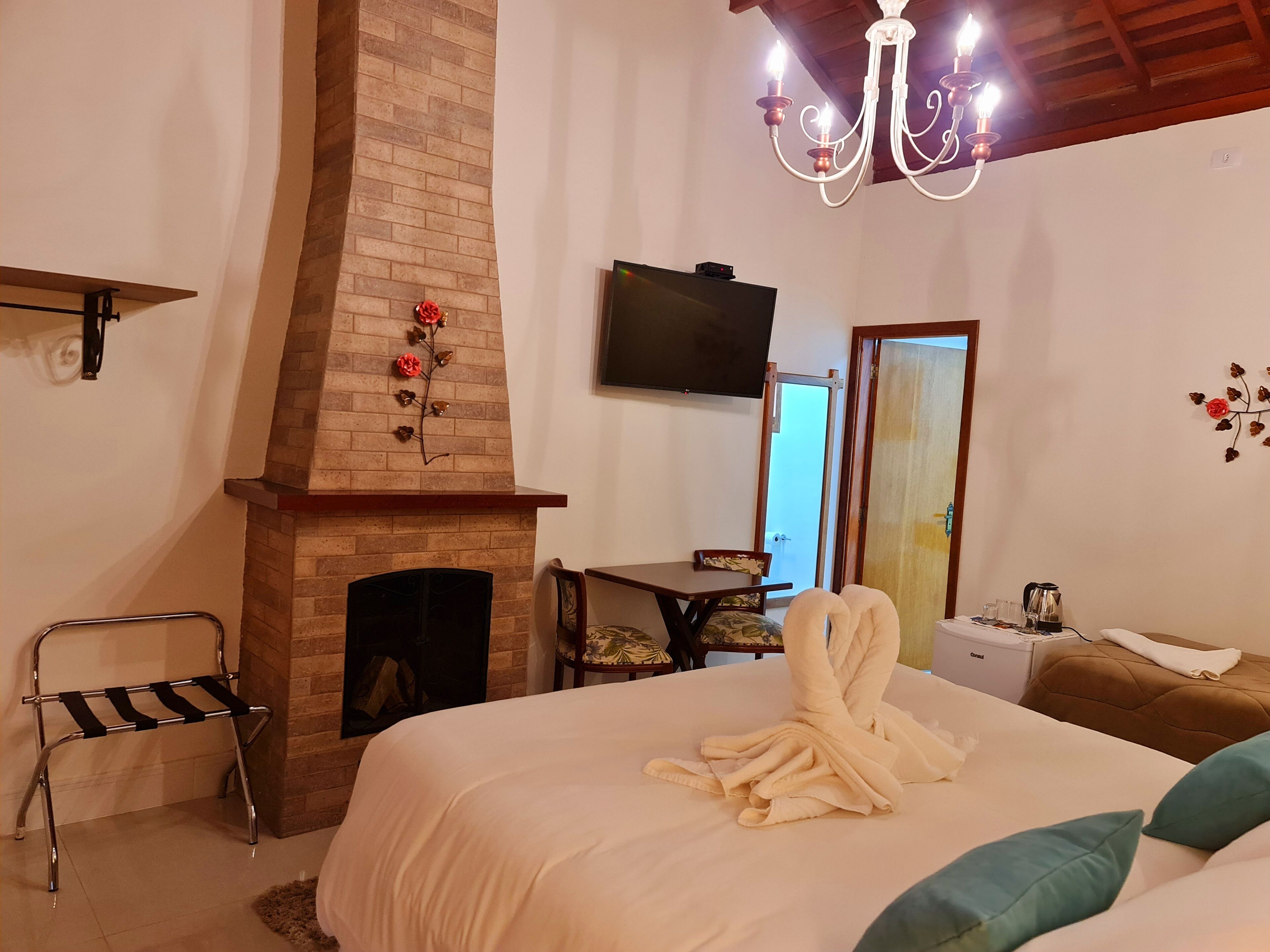 Un dormitorio con chimenea de ladrillo, televisor montado en la pared, cama con edredón blanco y candelabro.