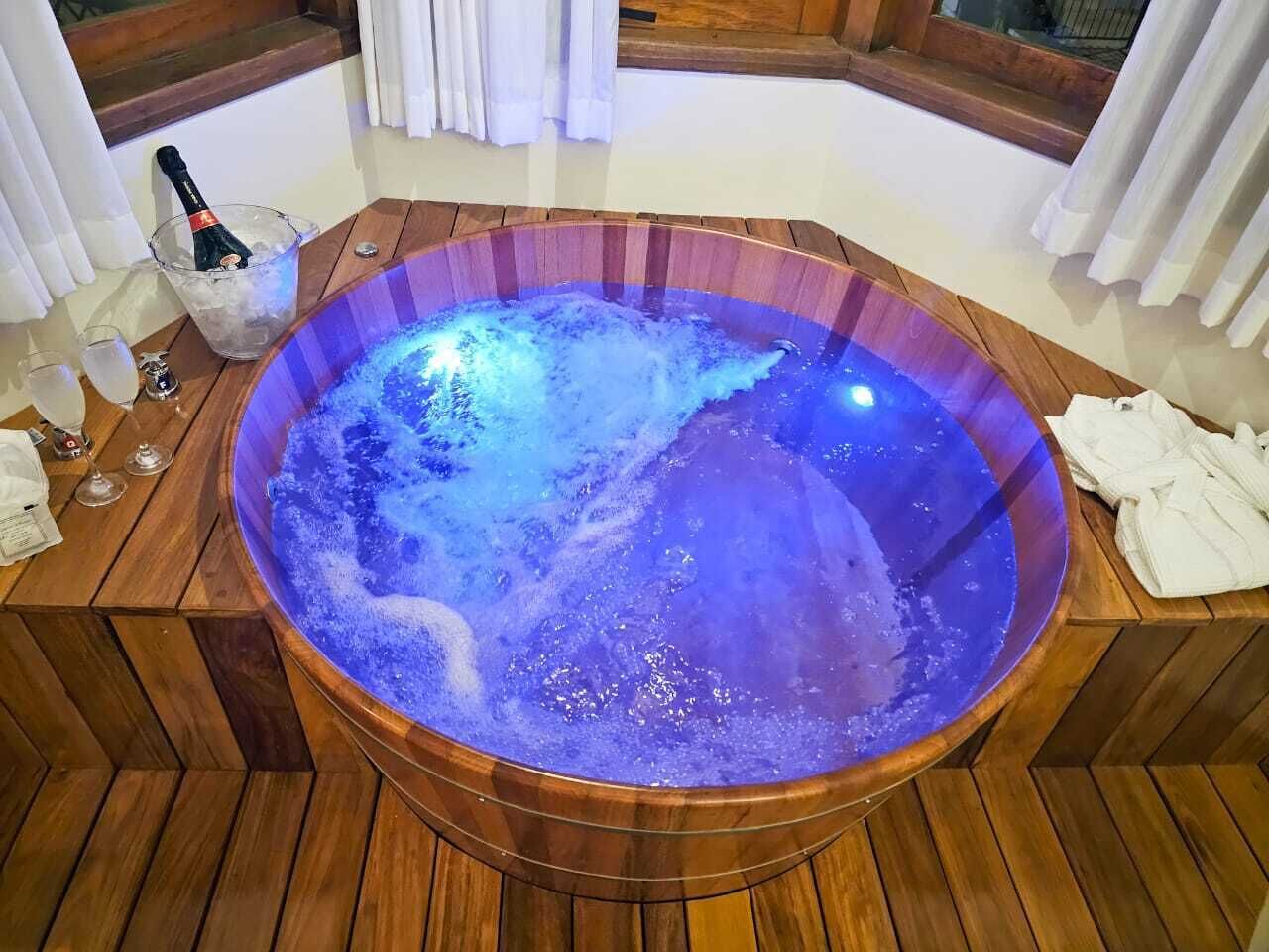 Een houten jacuzzi met blauw licht, omringd door houten vloerdelen, een champagnefles en glazen.