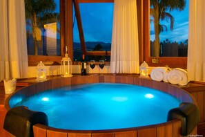 A hot tub with a view of palm trees and mountains, surrounded by wooden panels and lit by candles. - Pousada Valle das Flores (Camanducaia)