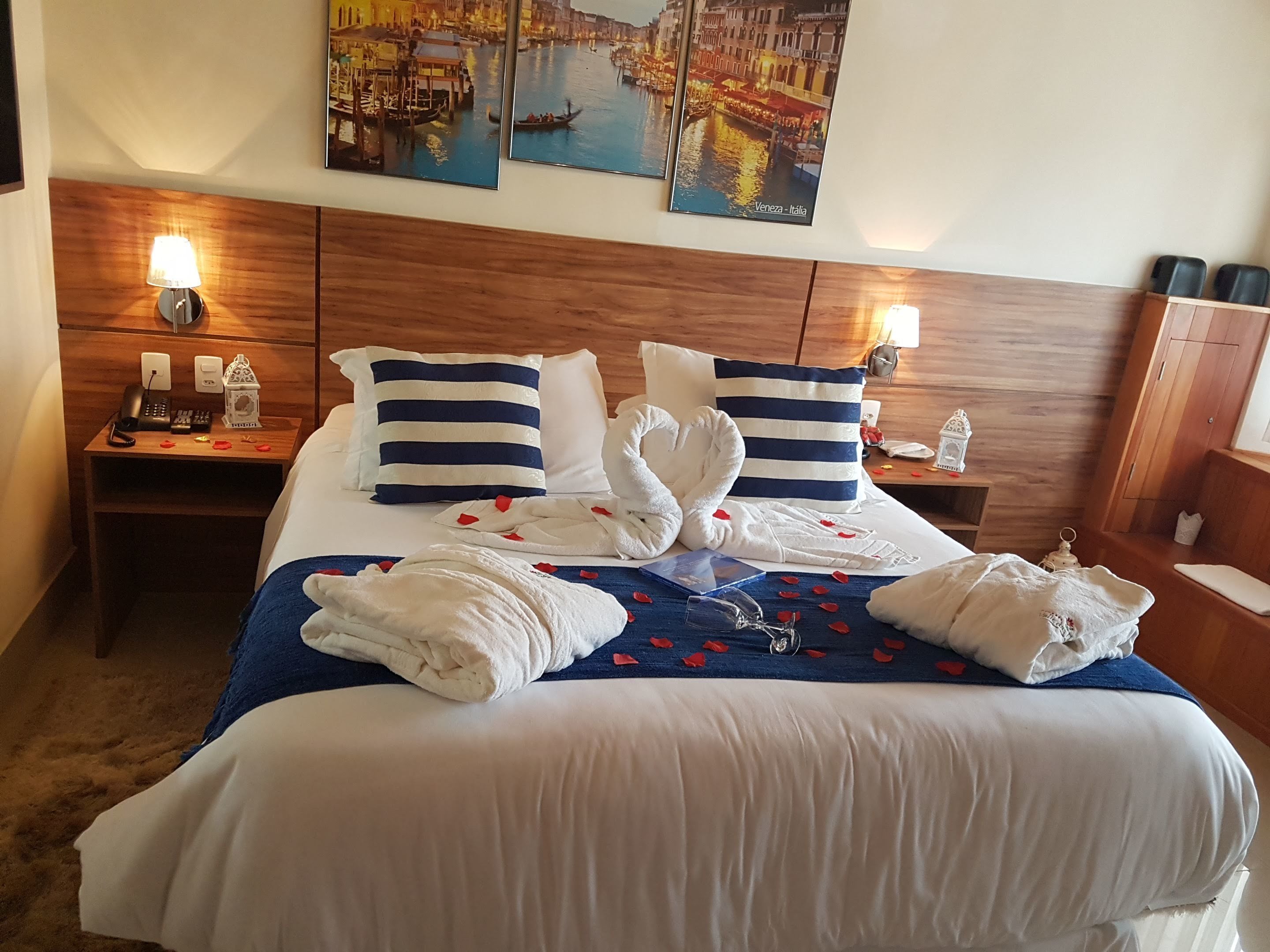 A hotel room with a large bed, two bedside tables, a wooden headboard, and wall art depicting a canal scene.