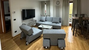 Una sala moderna con un sofá gris, un televisor de pantalla plana, piso de madera y un comedor con mesa y sillas.