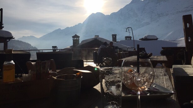 Una mesa con vasos y botellas, con vistas a un paisaje montañoso nevado y un cielo iluminado por el sol.