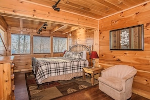 Ein Schlafzimmer einer Holzhütte mit einem Bett, einem Sessel, einem Nachttisch und einem Fenster mit Jalousien.