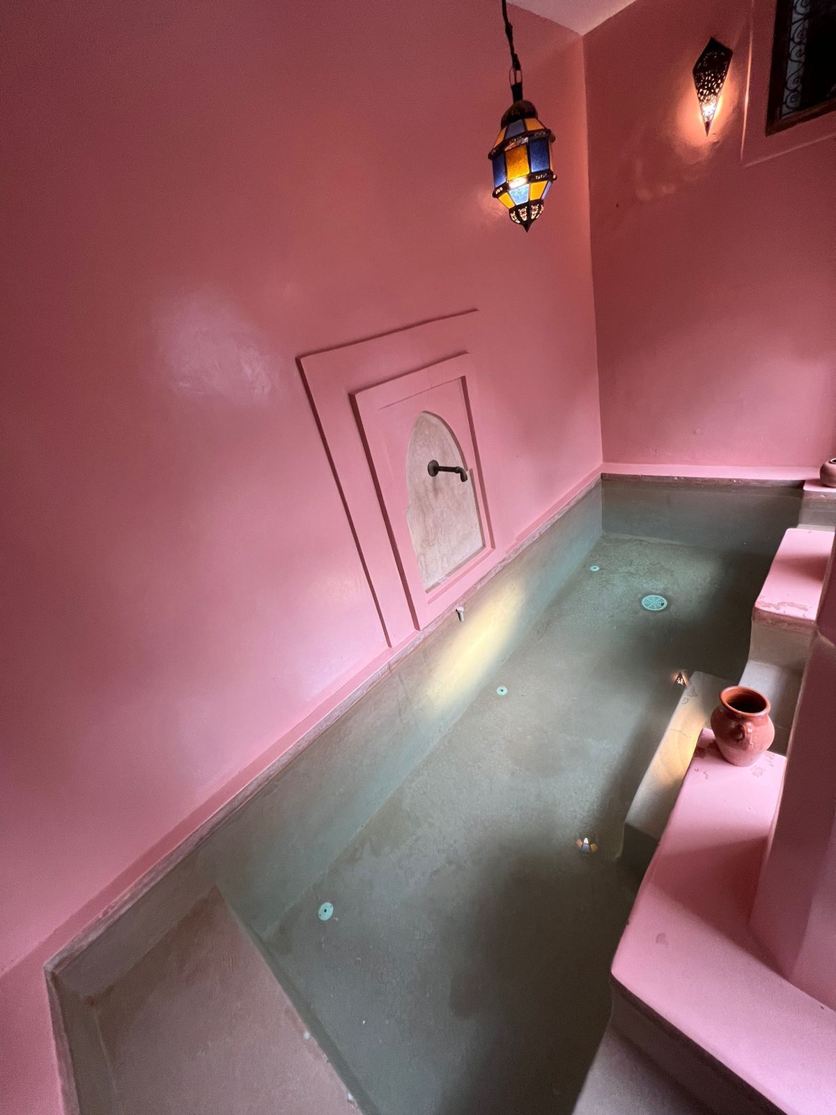 Ein Zimmer mit rosafarbenen Wänden, einer kleinen Tür, einem Fliesenboden und einer einzelnen Vase auf einer Ablagefläche.
