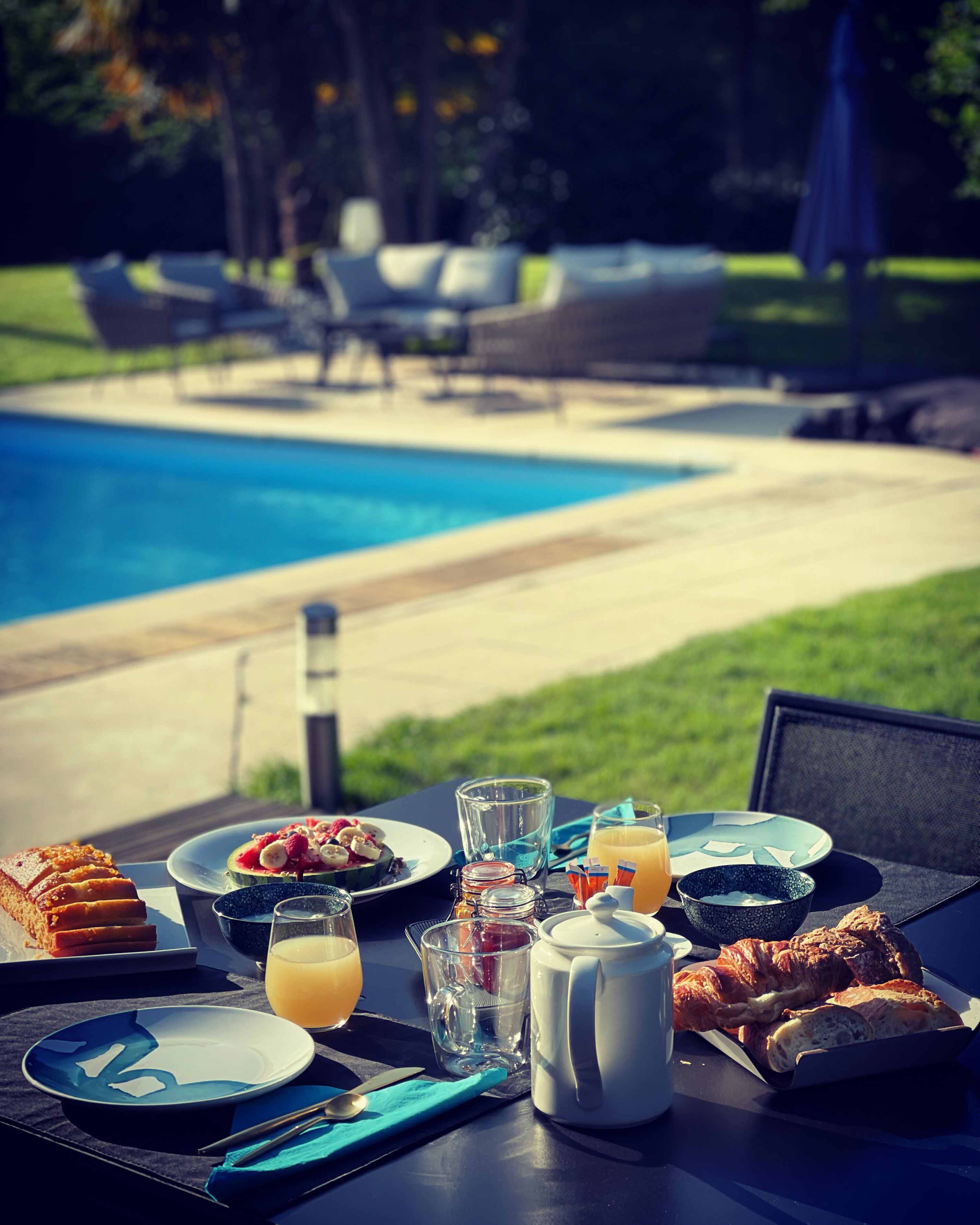 Ein Frühstückstisch mit Croissants, Obst und Saft an einem Pool mit Liegestühlen im Hintergrund.