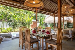 An outdoor dining area with wooden chairs and a glass table, surrounded by lush greenery and a thatched roof structure. - Renovated Villa Zayya, Central Oberoi Seminyak (Seminyak)