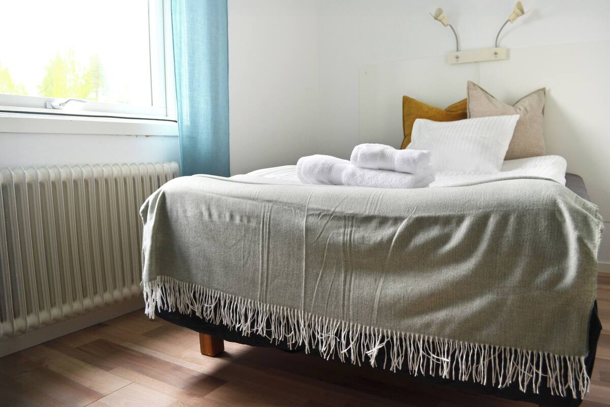 A neatly made bed with a white blanket and a fringed throw, a radiator, and a window with curtains.