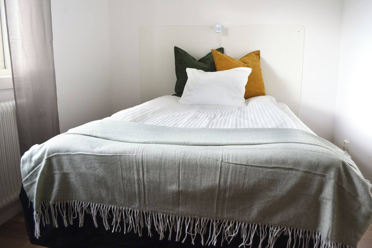 A neatly made bed with a white comforter and a grey blanket with fringe.