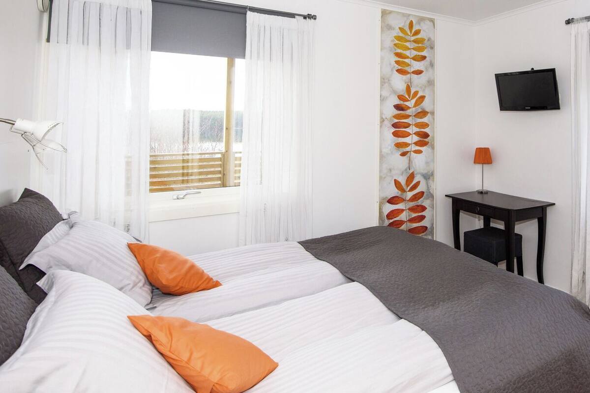 A double bed with white and gray bedding, two orange pillows, a small table with a lamp, and a TV on the wall.