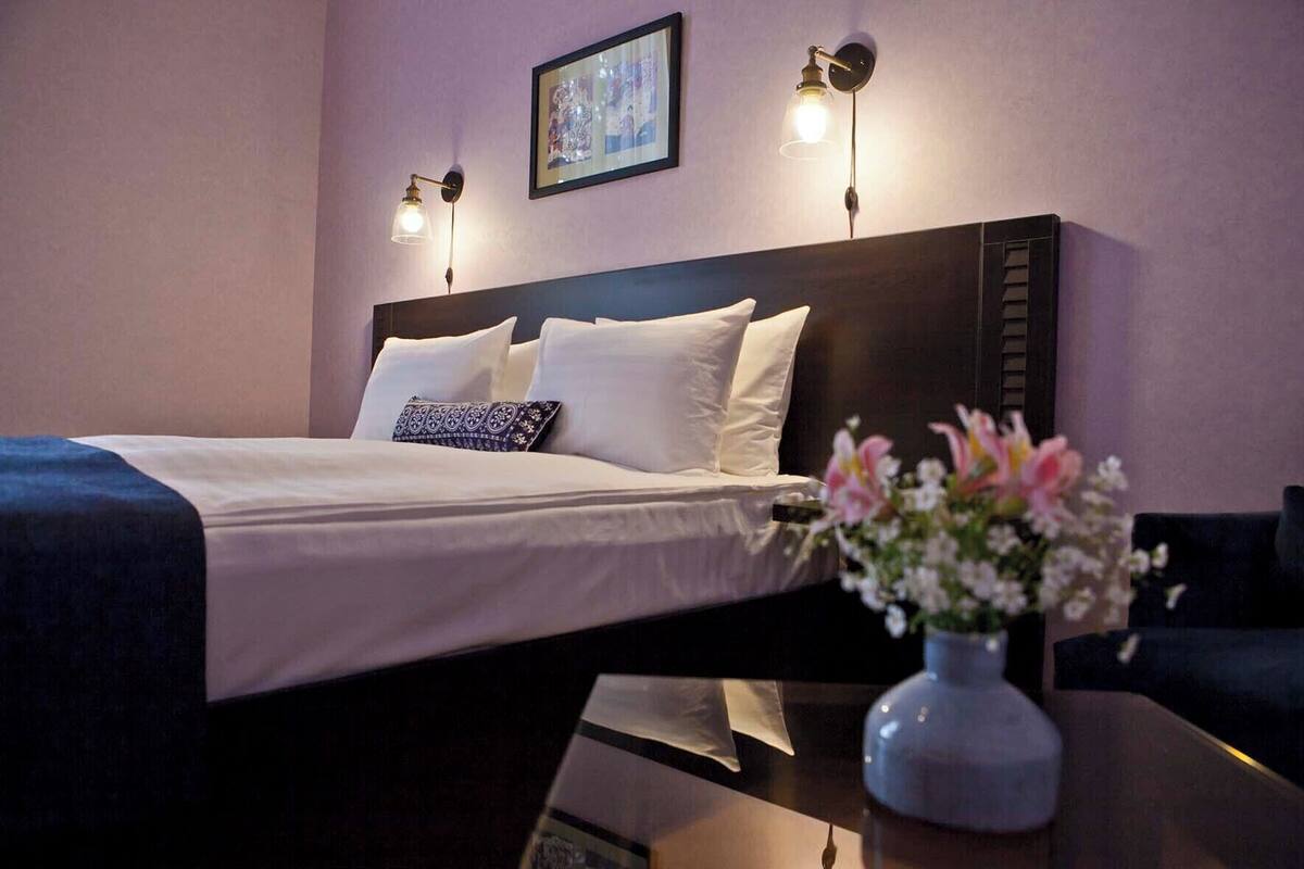 A neatly made bed with white linens and a patterned pillow, a dark headboard, and a vase with flowers on a table beside the bed.