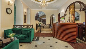 A lobby with a green sofa, a reception desk, a chandelier, and a staircase. - Hotel Bella Casa (Tbilisi)