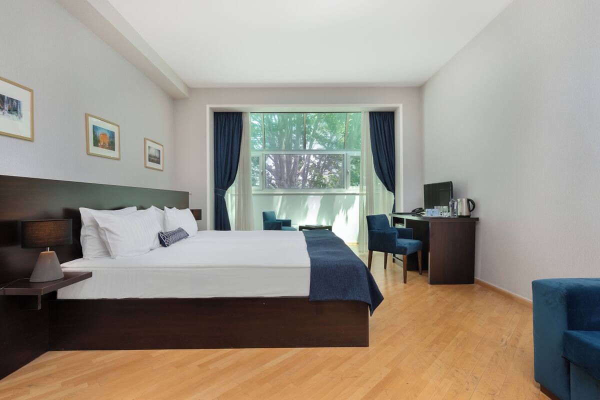 A hotel room with a large bed, a desk, a chair, and a view of trees through the window.