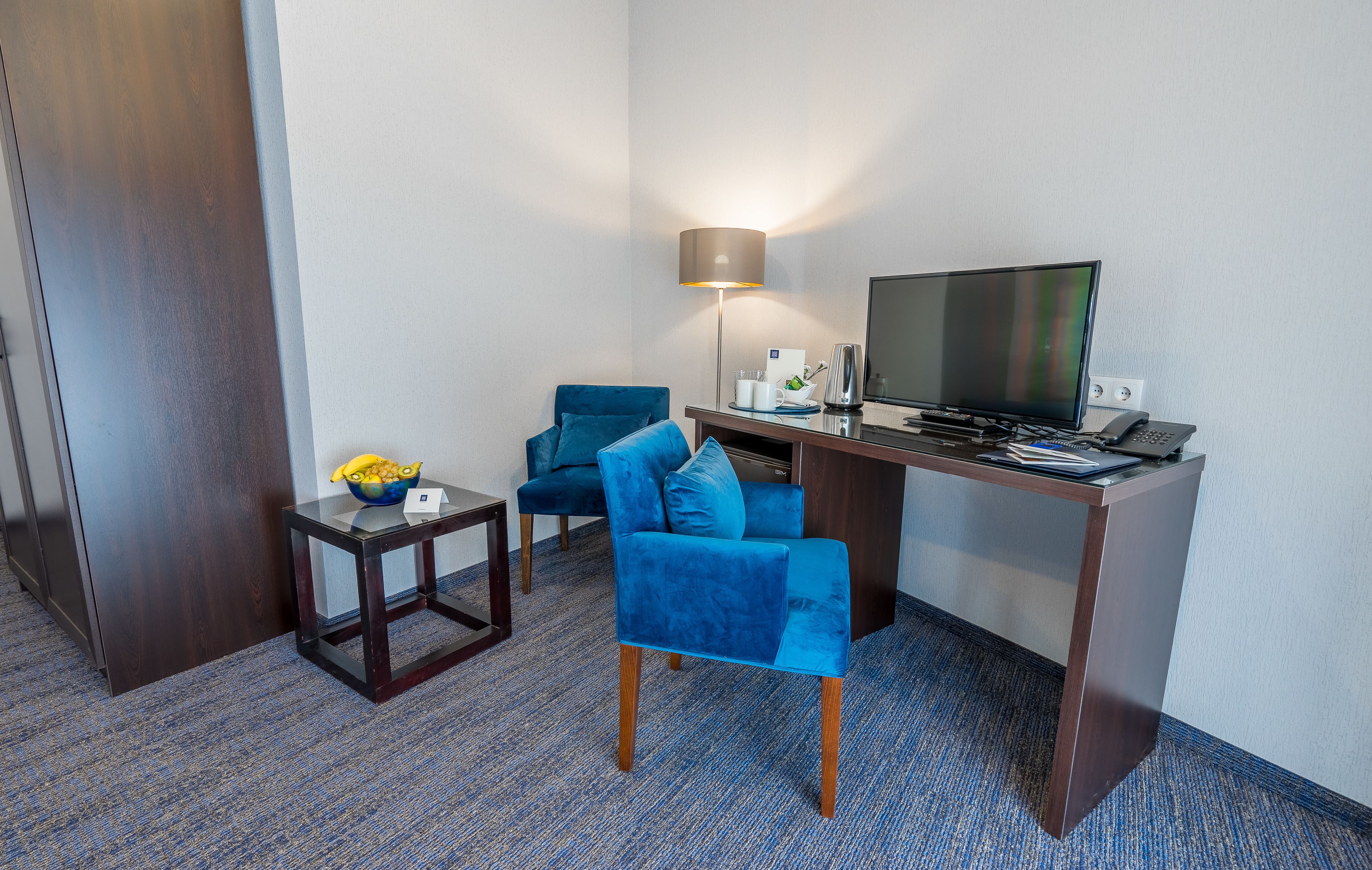 Une chambre d’hôtel avec un bureau, une chaise bleue, une petite table sur laquelle se trouve un saladier de fruits, une lampe et un téléviseur.