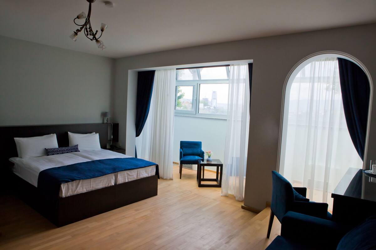 A hotel room with a bed, a chair, a small table, and a view of the city through large windows.