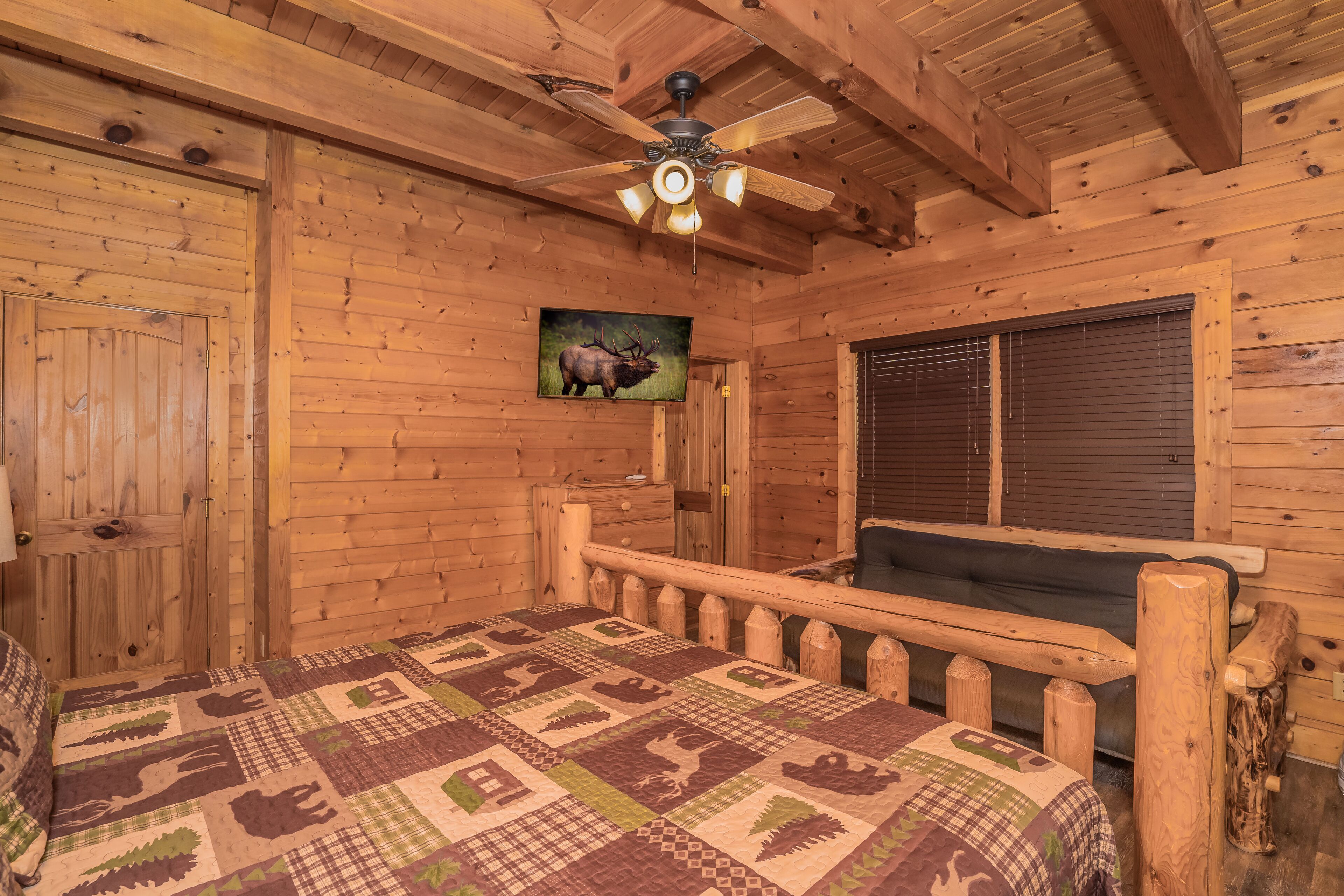 A wooden cabin room with a bed, a ceiling fan, a window with blinds, and a wall-mounted picture of an elk.
