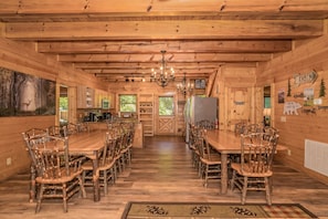 A rustic cabin interior with a long wooden dining table and chairs, a chandelier, and a forest-themed painting on the wall. - Creekside Getaway: Top Amenities, Game Room, Fire Pit area on the creek, Ample Parking, Gatlinburg! (Pigeon Forge)