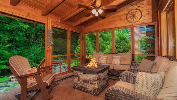 A wooden interior with a stone fire pit, wicker furniture, and large windows overlooking a forest. - Creekside Getaway: Top Amenities, Game Room, Fire Pit area on the creek, Ample Parking, Gatlinburg! (Pigeon Forge)