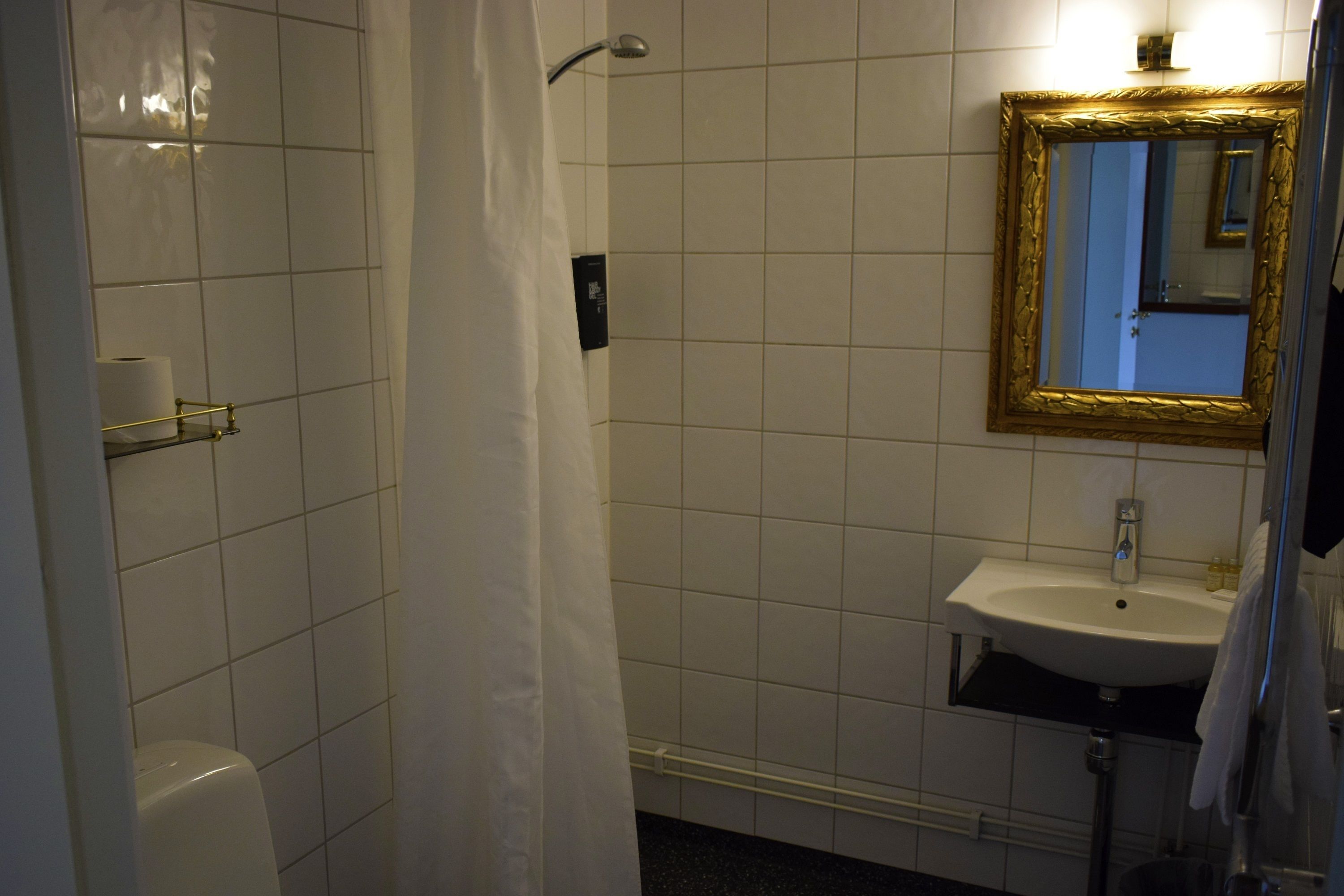 Un baño con azulejos blancos, un espejo con marco dorado, un lavabo y una cortina de ducha.
