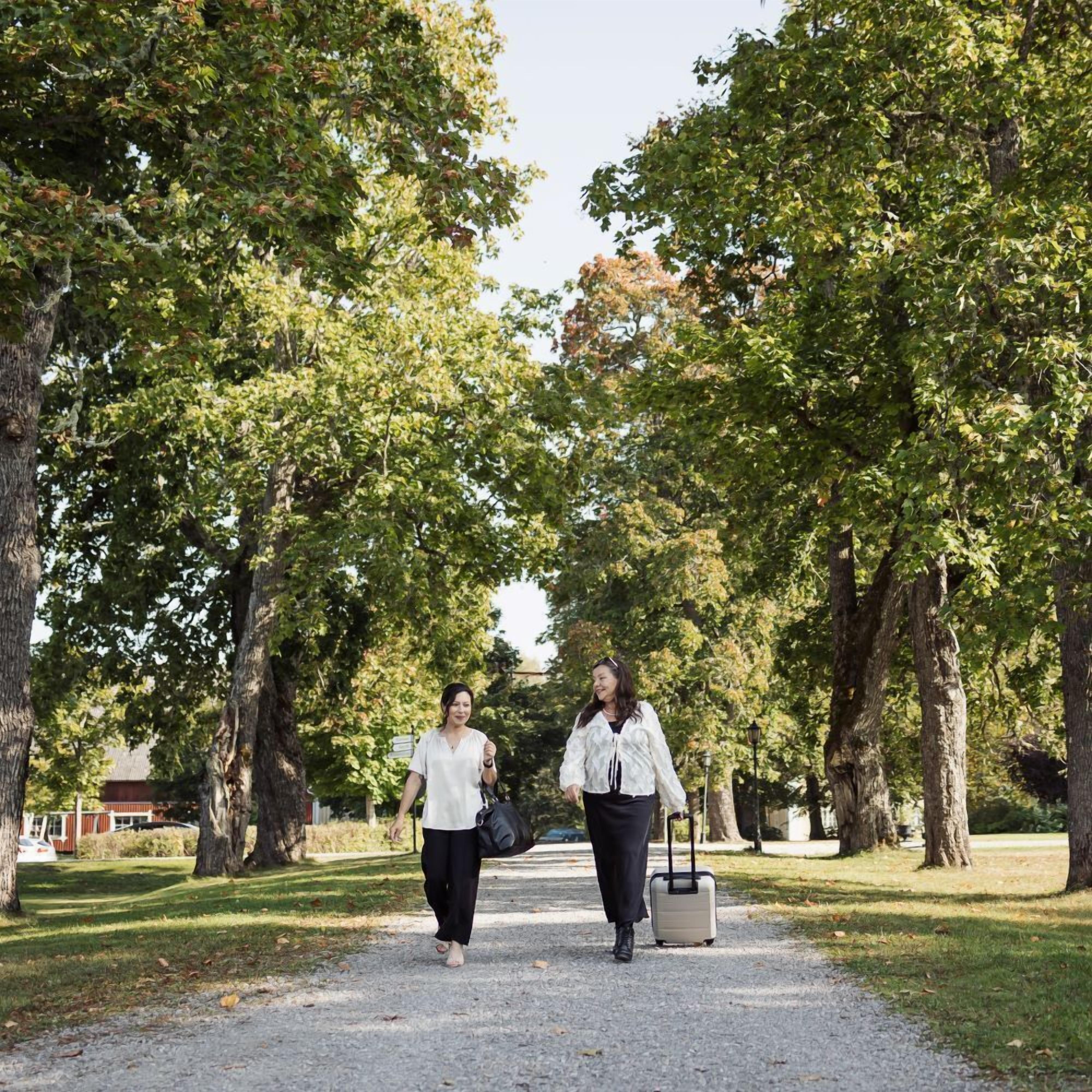 Två personer promenerar på en grusgång i en park, med träd på båda sidor.