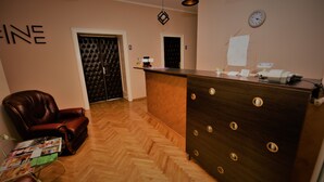 Uma área de recepção com piso de madeira em padrão herringbone, uma poltrona de couro, um balcão de recepção com porta-papéis e um relógio na parede.