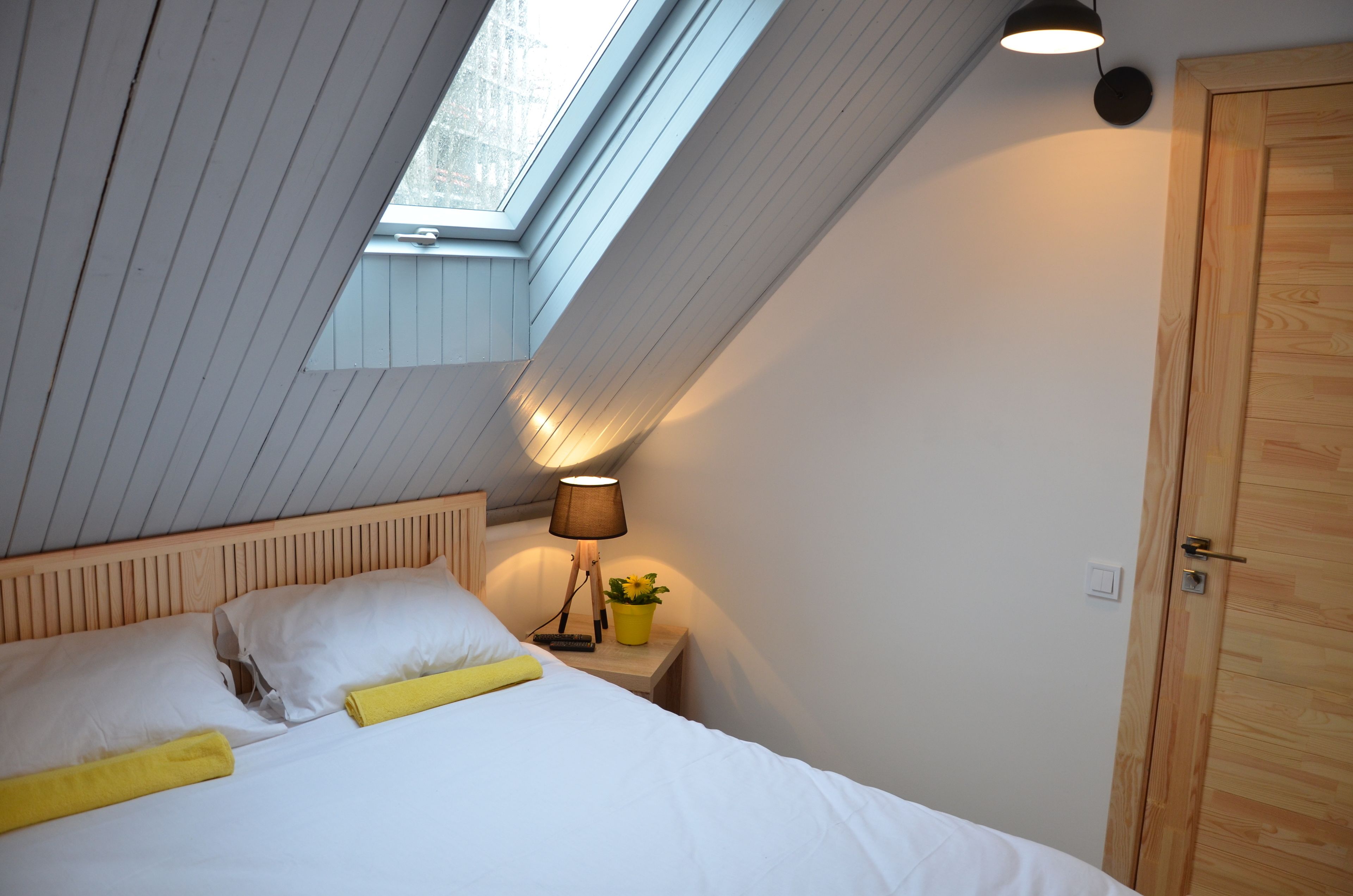 Ein Schlafzimmer mit schrägen Wänden, einem Bett mit weißer Bettwäsche, einer Nachttischlampe und einer Holztür.