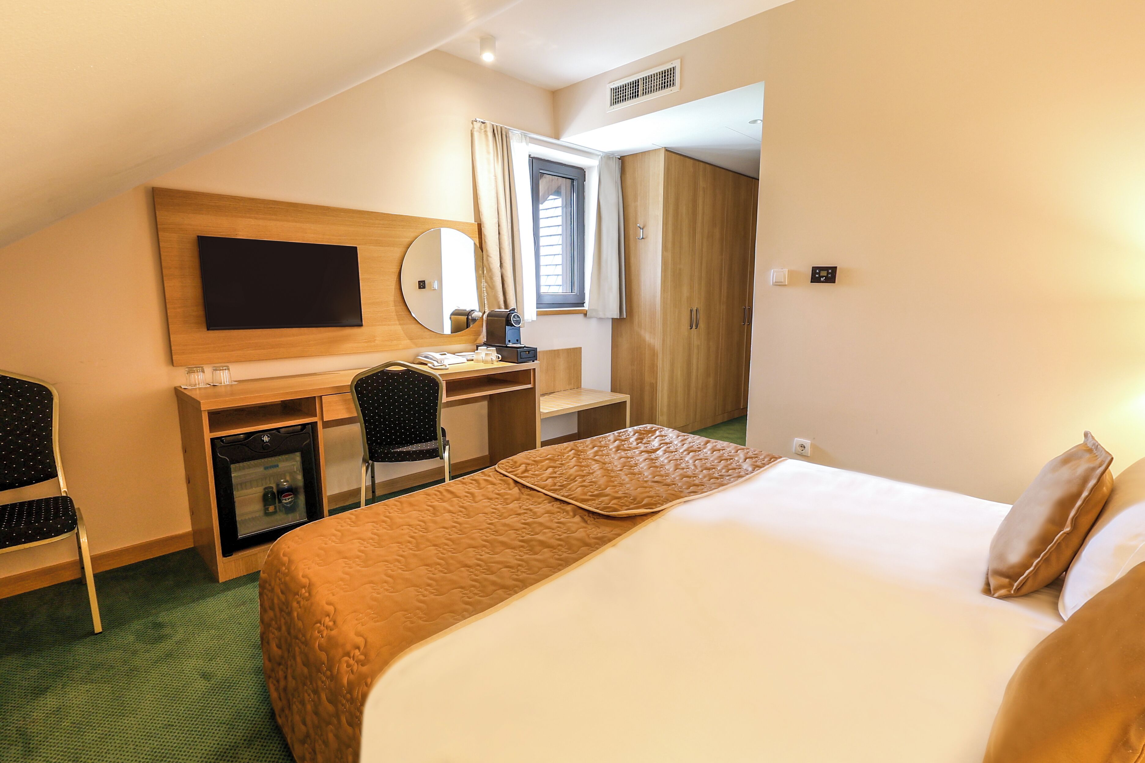 A hotel room with a bed, a desk with a computer, a TV, and a minibar.