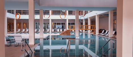 Une vaste piscine intérieure à plusieurs niveaux, dotée de cloisons en verre transparent et d’un escalier central.
