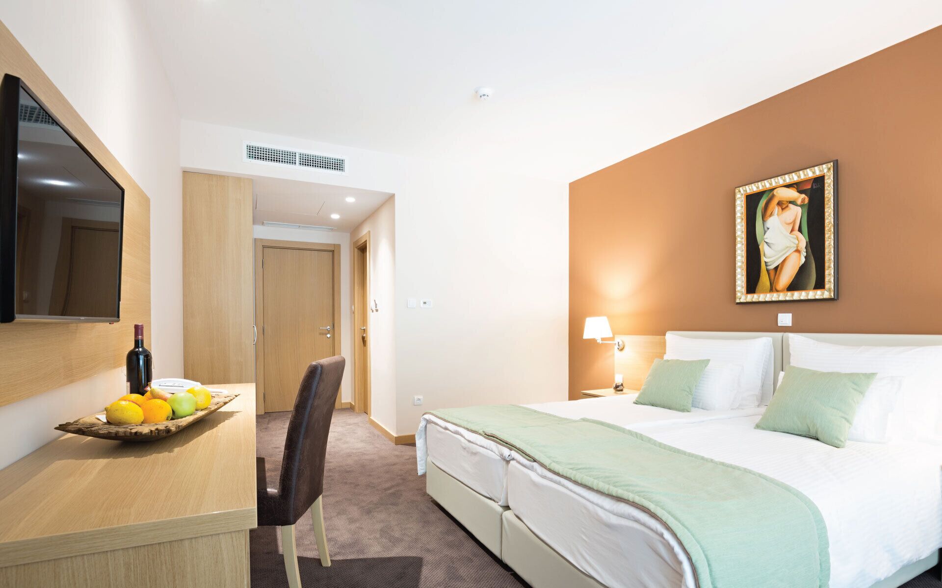 A hotel room with a bed, a desk with a fruit bowl, a chair, a TV, and a painting on the wall.