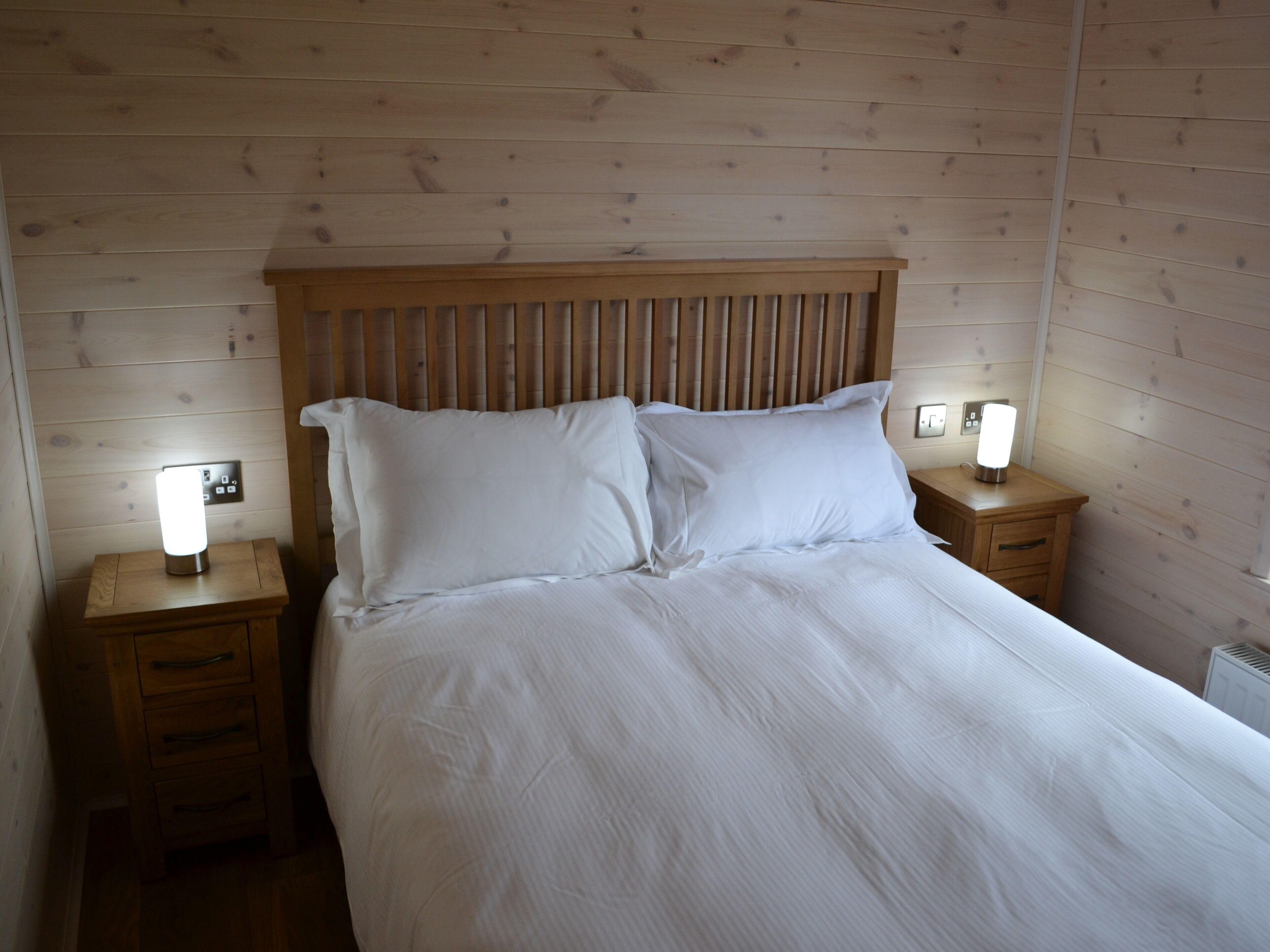 A neatly made bed with white linens in a room with wooden panel walls. There are two bedside tables, each with a lamp and a power outlet.
