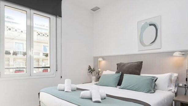 A modern bedroom with a large bed, a view of a building through a window, and a simple decor. - One Bedroom Apartment with balcony (Barcelona)