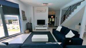 A modern living room with a dark blue sofa, a white coffee table, and a flat-screen TV on a white stand. There is a staircase to the right and a view of the outdoors through large windows. - Villa Palmela in Serra da Arrábida (Palmela)