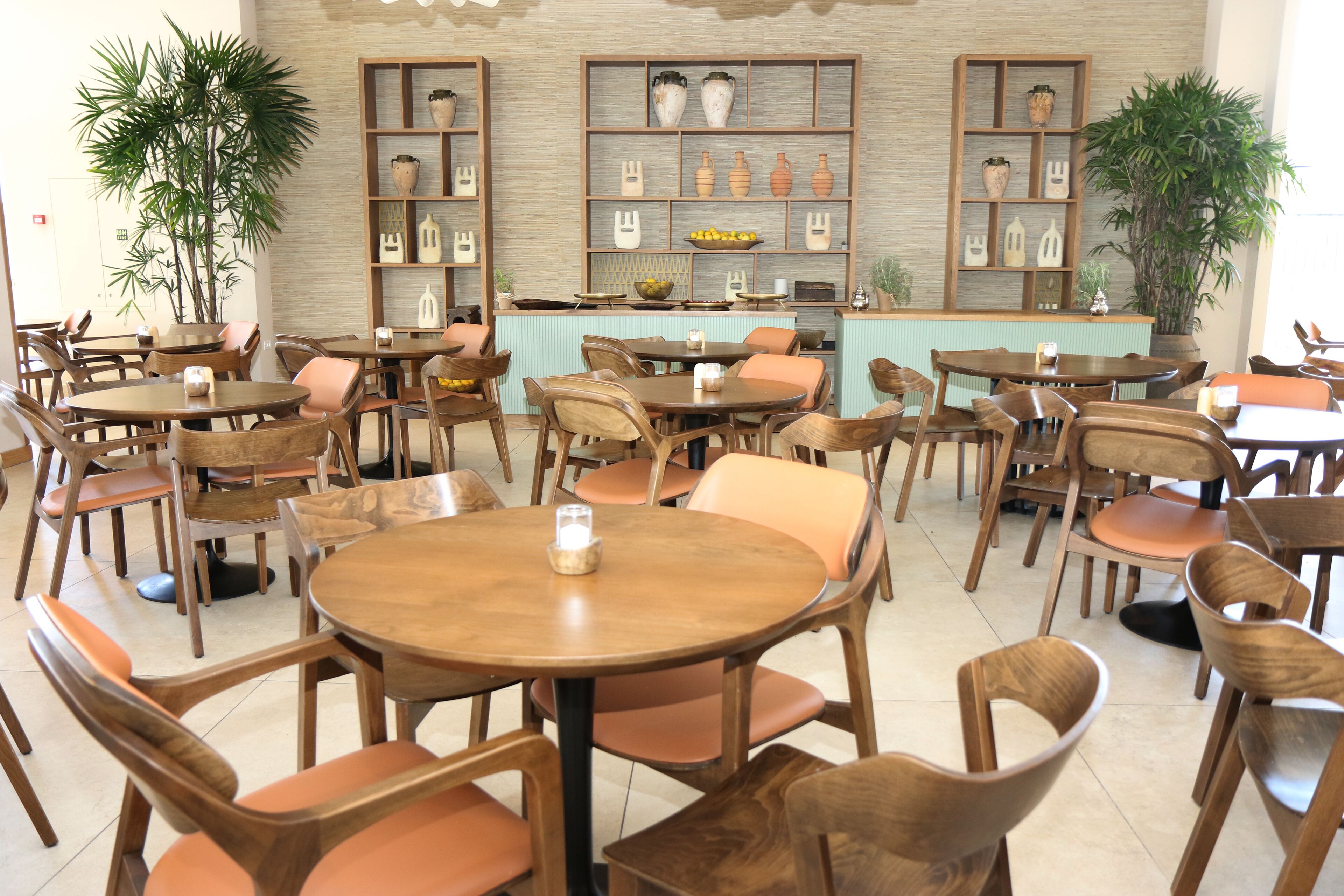 Un restaurant avec des tables rondes et des chaises en bois, un comptoir à la façade bleu clair, et des étagères ornées de poteries et de plantes.
