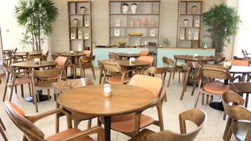 Ein Restaurant mit runden Tischen und Holzstühlen, eine Theke mit hellblauer Front sowie Regale mit Keramik und Pflanzen.
