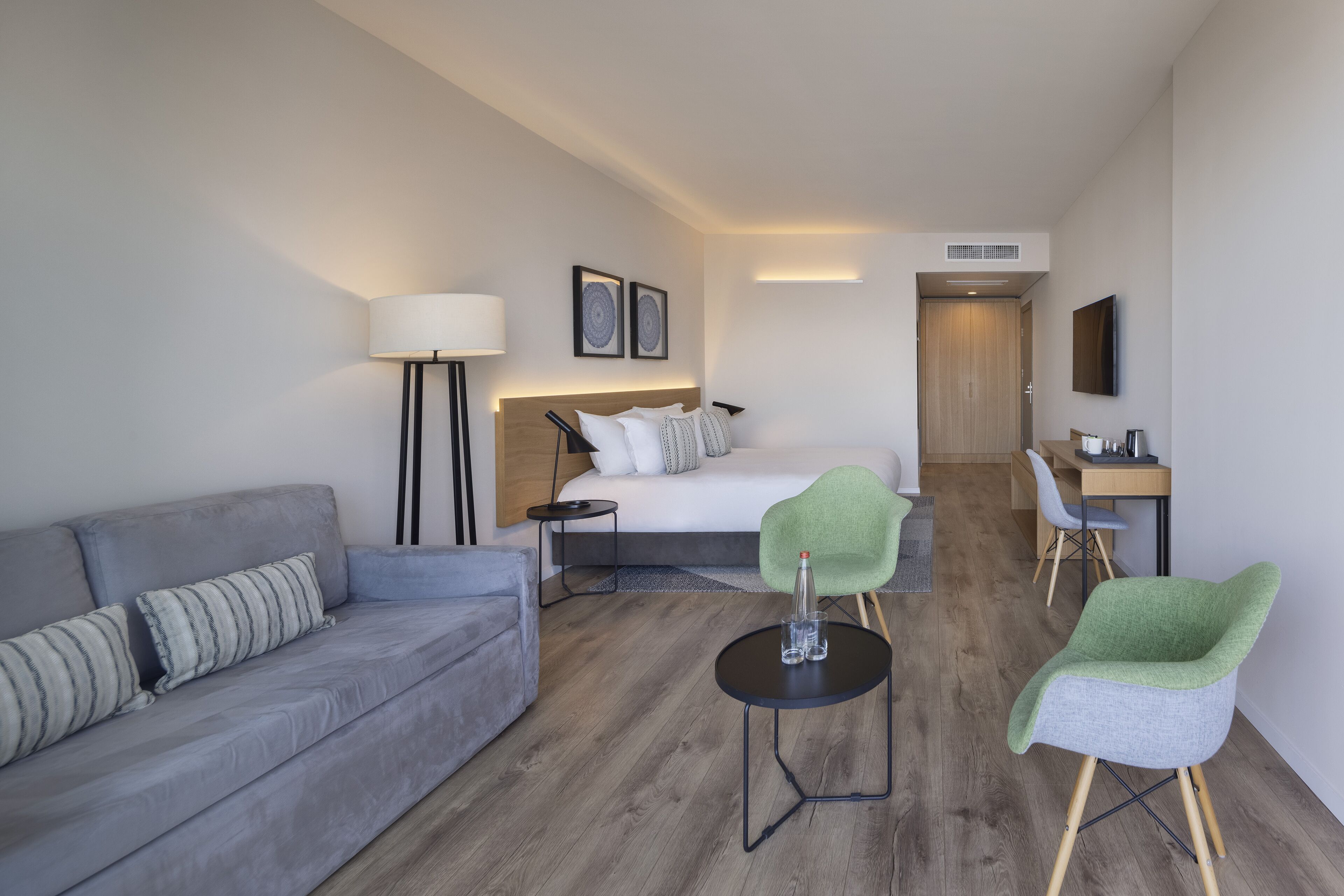 A modern hotel room with a grey sofa, a bed with white bedding, a green chair, a small round table with a bottle and glasses, and a desk with a chair.