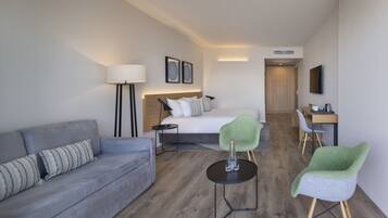 A modern hotel room with a grey sofa, a bed with white bedding, a green chair, a small round table with a bottle and glasses, and a desk with a chair.