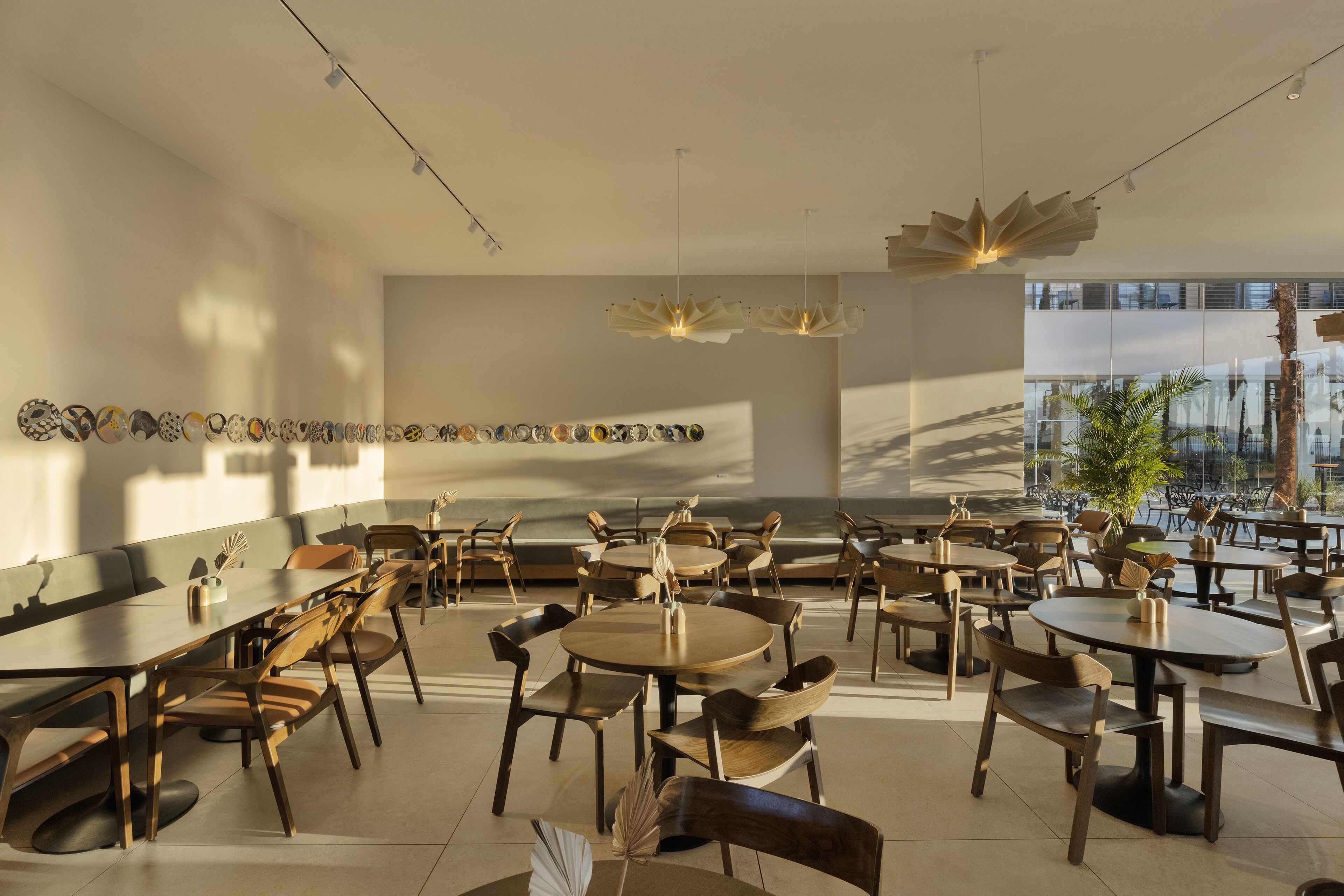 A modern dining area with multiple tables and chairs, a long wall of decorative plates, and a large window with a view of palm trees.