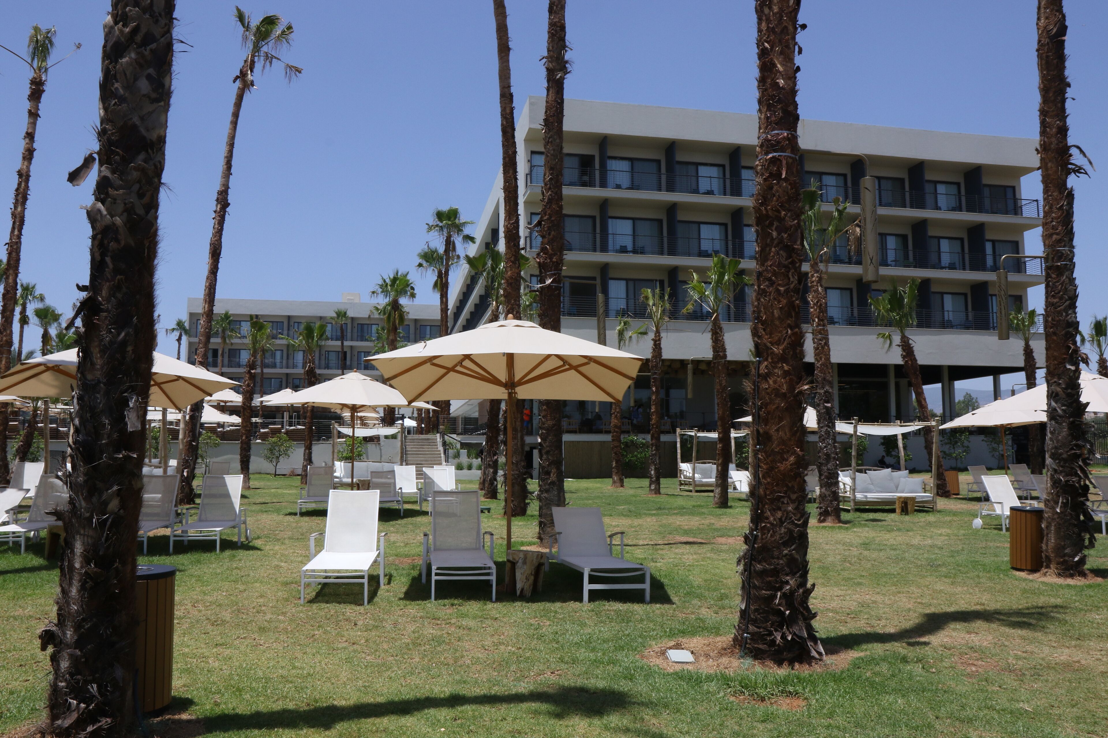 Ein Resort mit mehreren Liegestühlen und Sonnenschirmen, Palmen und einem modernen Gebäude mit Balkonen.
