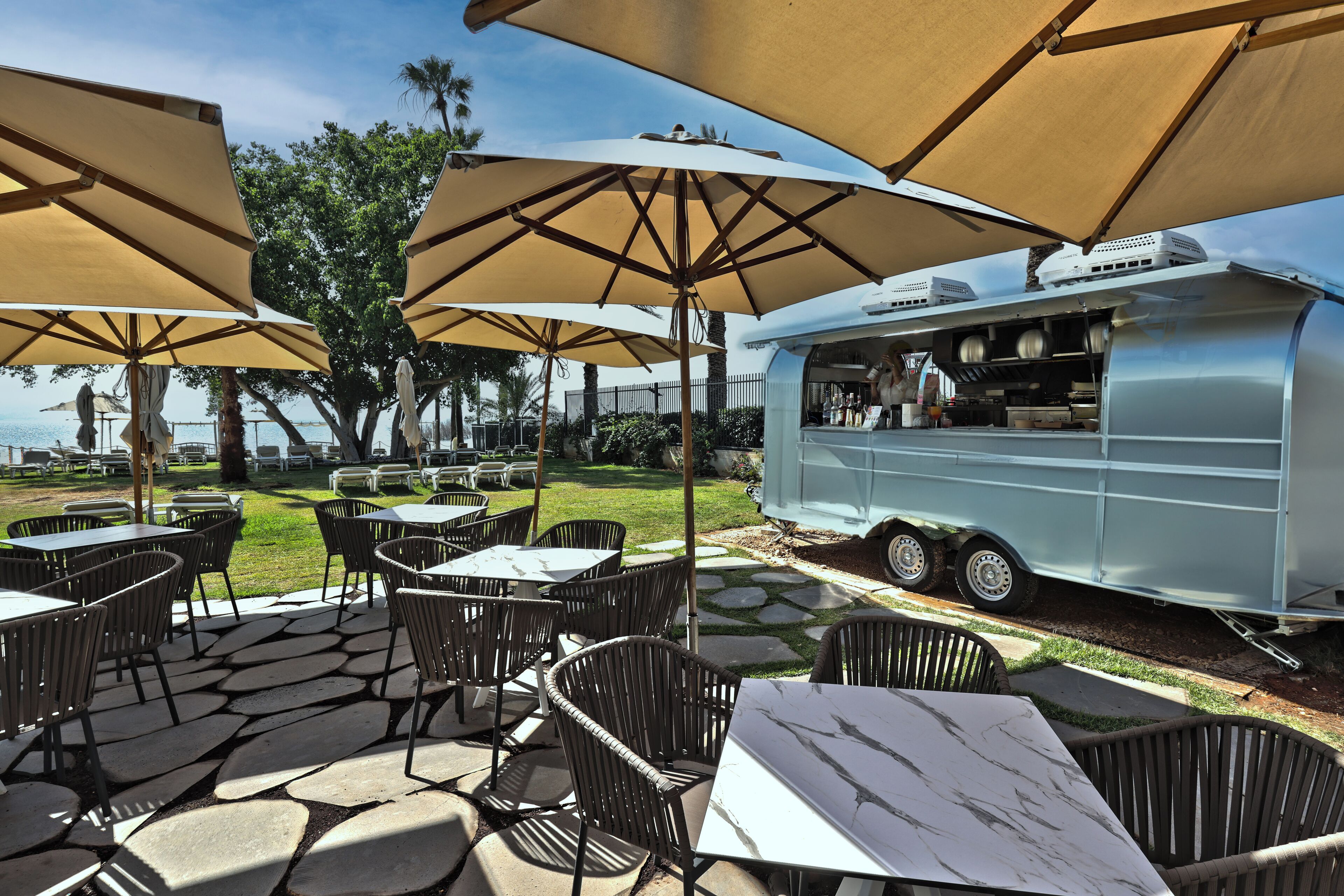 Een terras buiten met tafels en stoelen onder grote parasols, een foodtruck en uitzicht op de oceaan.