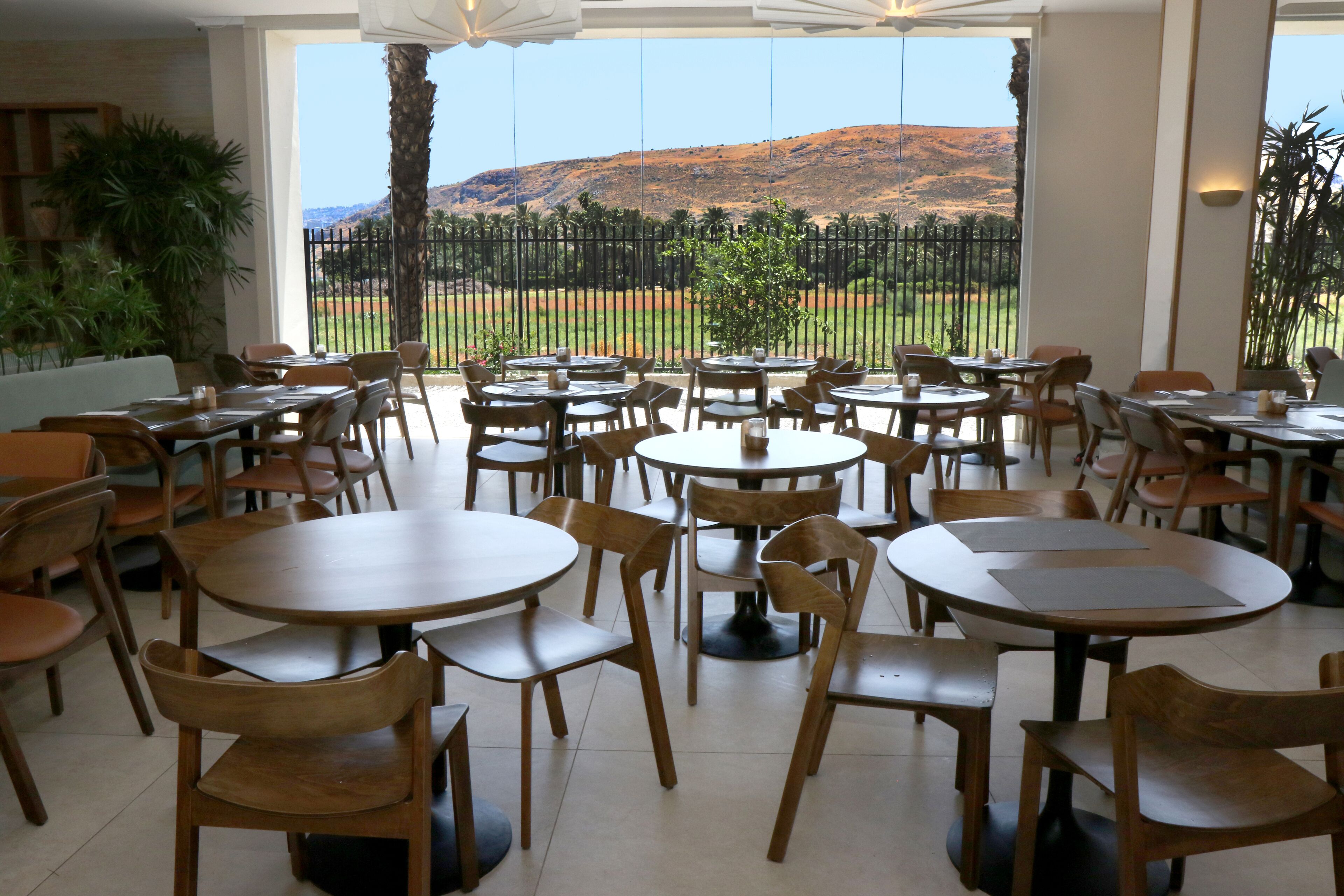 Een restaurant met ronde tafels en houten stoelen, grote ramen met uitzicht op een glooiend landschap, en een zwarte metalen hek op de achtergrond.