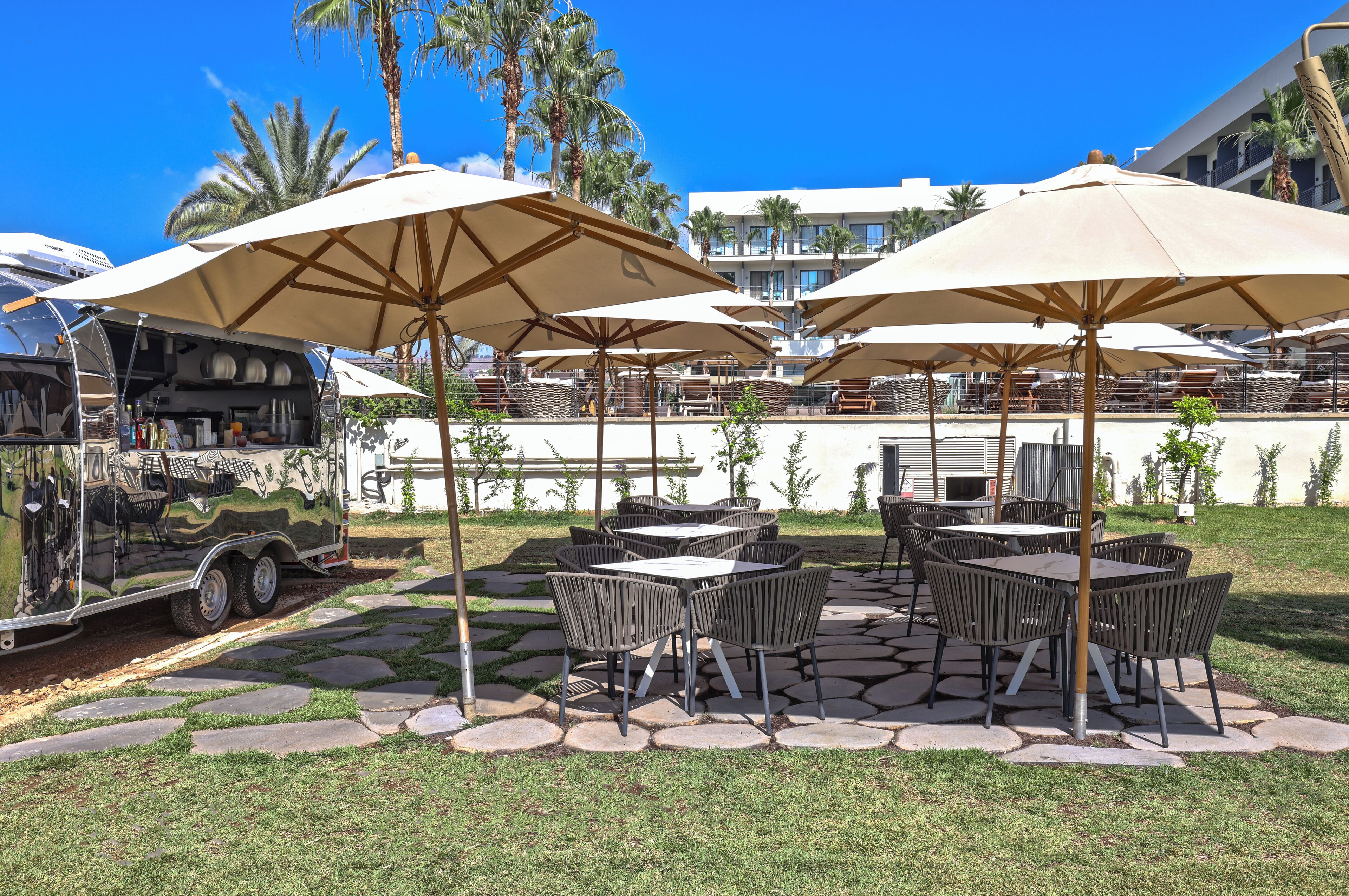 Een terras buiten met tafels en stoelen onder grote parasols, een barbecue en een zwembad met palmbomen en een gebouw op de achtergrond.