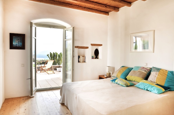 A bedroom with a large bed, a wooden ceiling, a glass door leading outside, and a small table with a lamp.